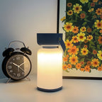 Clérachic cordless cylinder lamp with a blue handle glowing warm on a pale surface beside a black alarm clock and a floral print.