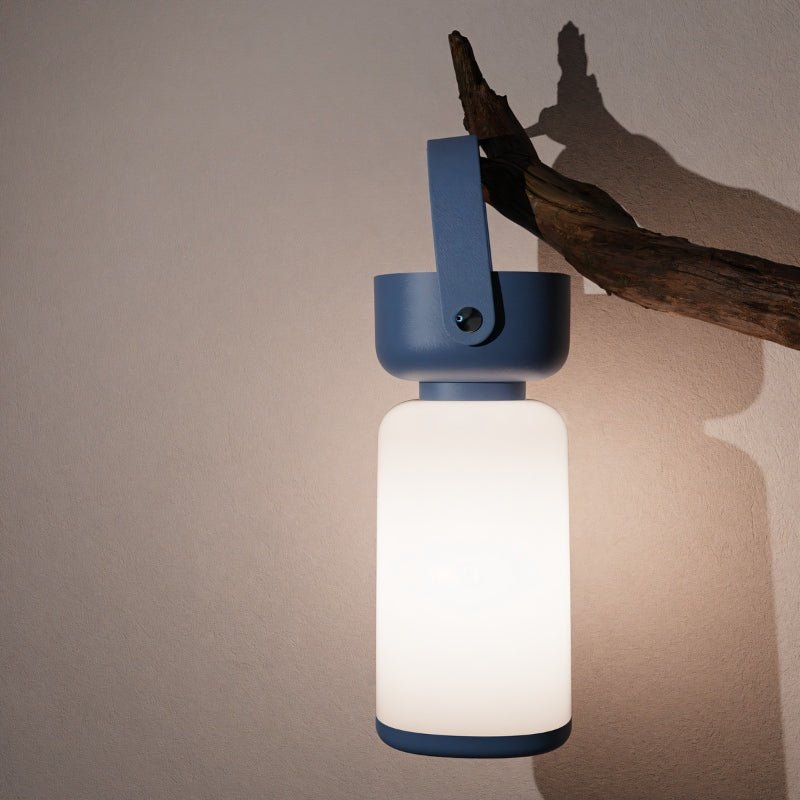Clérachic cordless cylinder lamp with a blue handle hung from a driftwood branch against a plain wall, glowing softly.