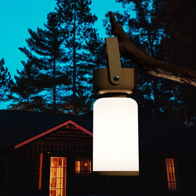 Clérachic cordless cylinder lamp with a brown handle hanging from a branch at night, a lit cabin and pines behind it.