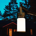 Clérachic cordless cylinder lamp with a brown handle hanging from a branch at night, a lit cabin and pines behind it.
