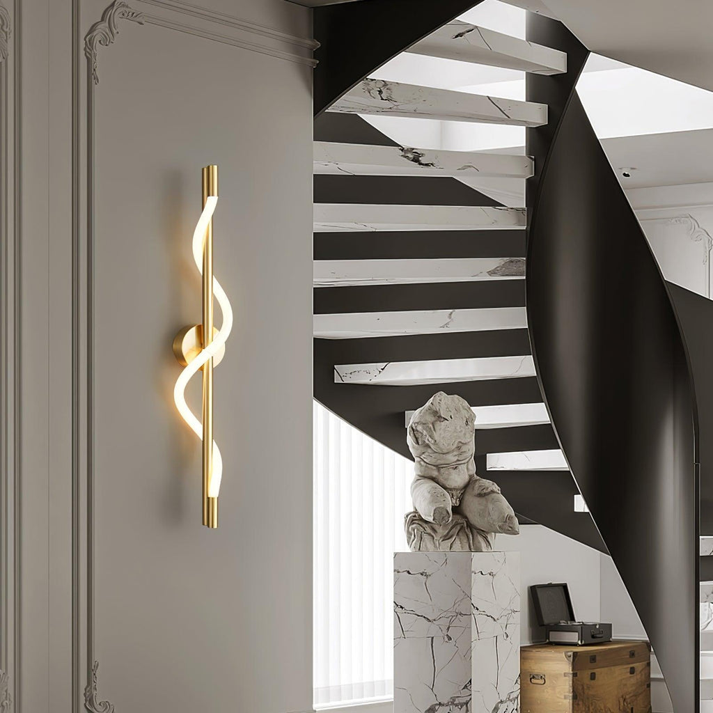A brass Clérachic wall light with a lit spiral band glows on a pale wall beside a black curving staircase and a marble plinth.