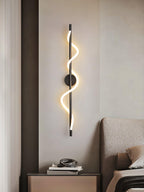 Above a bedside table in a beige bedroom, a black Clérachic wall light glows with a lit spiral band by a padded headboard.