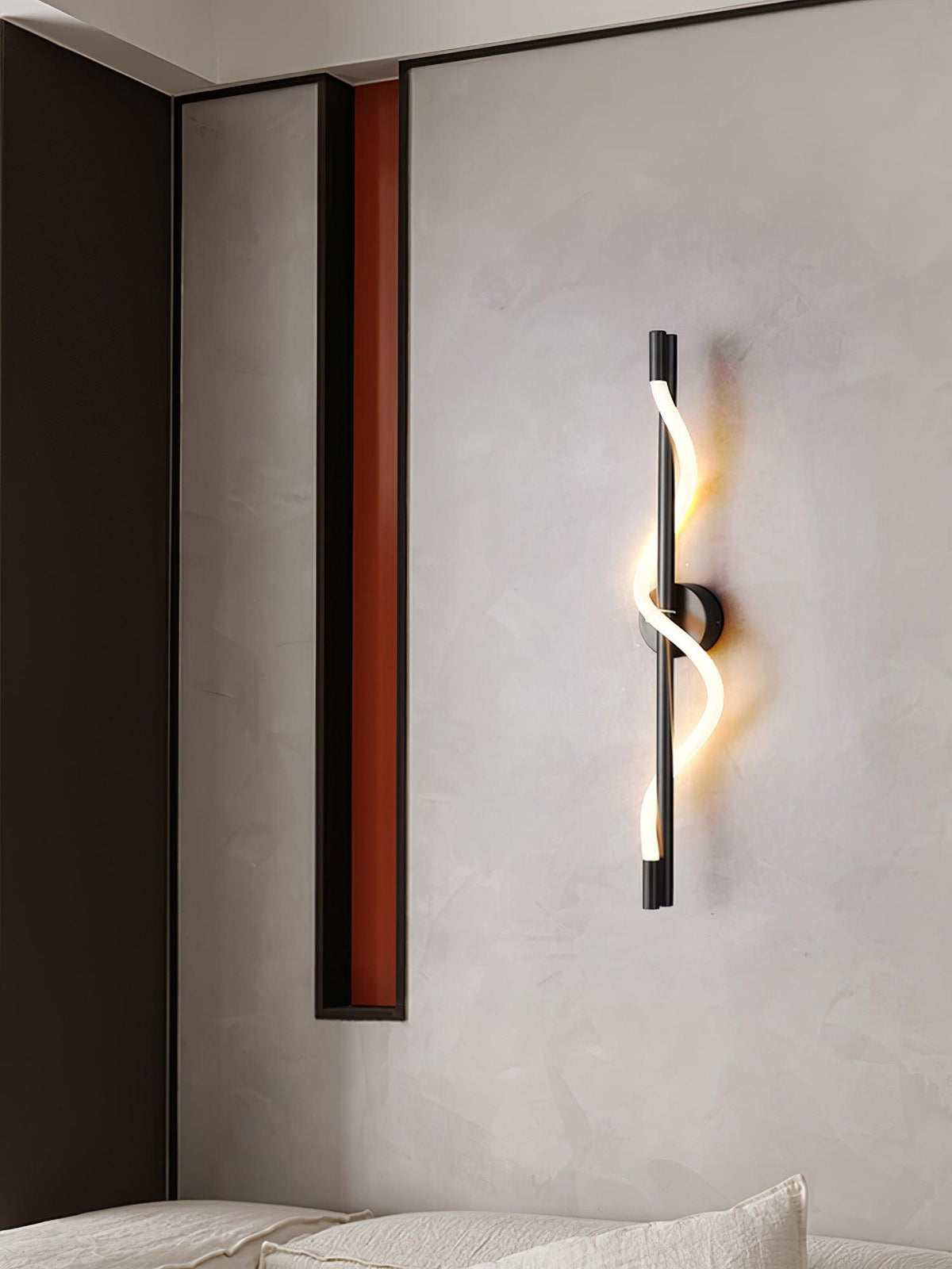 On a pale wall, a black Clérachic wall light with a lit twisted band stands out, with a dark red recess and bed at the lower left.