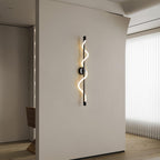 A slim black Clérachic wall light with a lit twisting band on a cream wall beside a window with vertical blinds.
