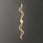 A long brass Clérachic wall light with a two-turn lit spiral, mounted on a dark grey wall that fades to black.