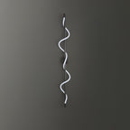 A black Clérachic wall light with a cool white spiral band on a dark grey wall, with the round backplate behind the rod.