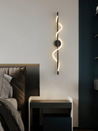 Over a dark bedside table, a black Clérachic wall light with a lit spiral band hangs by a deep blue panel.