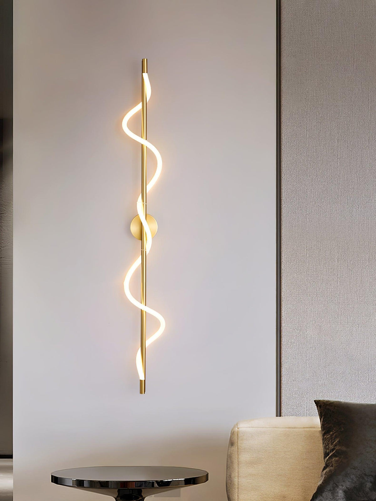 A brass Clérachic wall light with a lit two-turn spiral hangs on a pale wall above a round side table and a sofa.