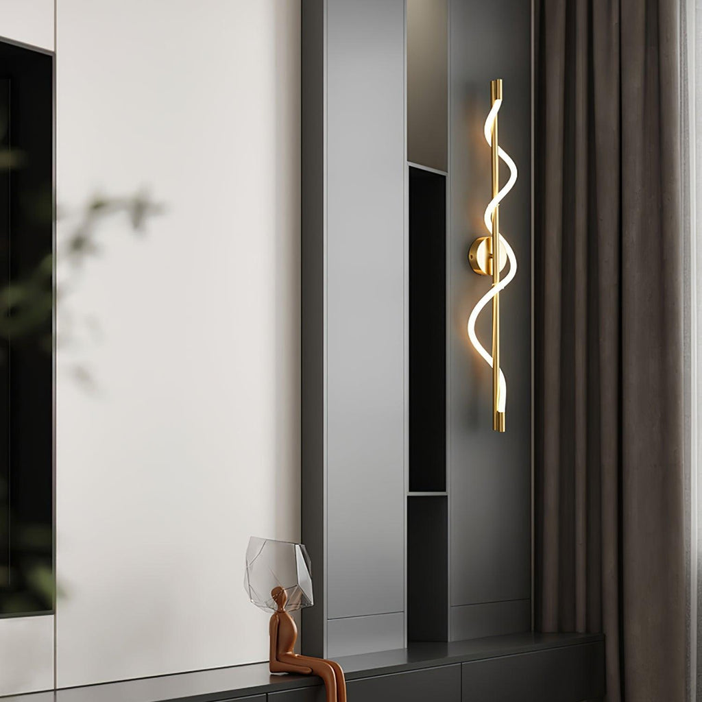 A brass Clérachic wall light with a lit spiral band hangs by dark shelving and a curtain, with a small sculpture below.