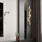 A brass Clérachic wall light with a lit spiral band hangs by dark shelving and a curtain, with a small sculpture below.