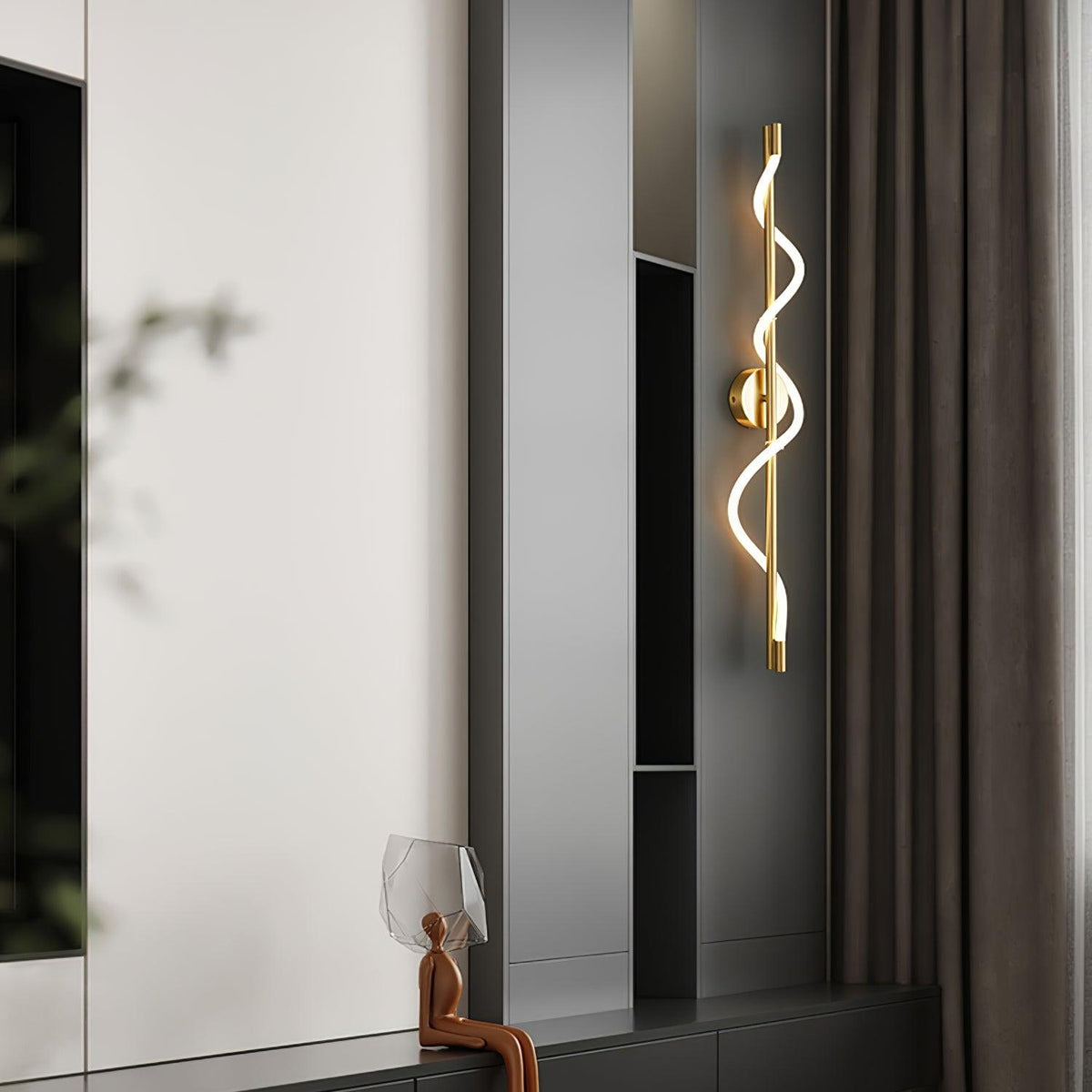 A brass Clérachic wall light with a lit spiral band hangs by dark shelving and a curtain, with a small sculpture below.