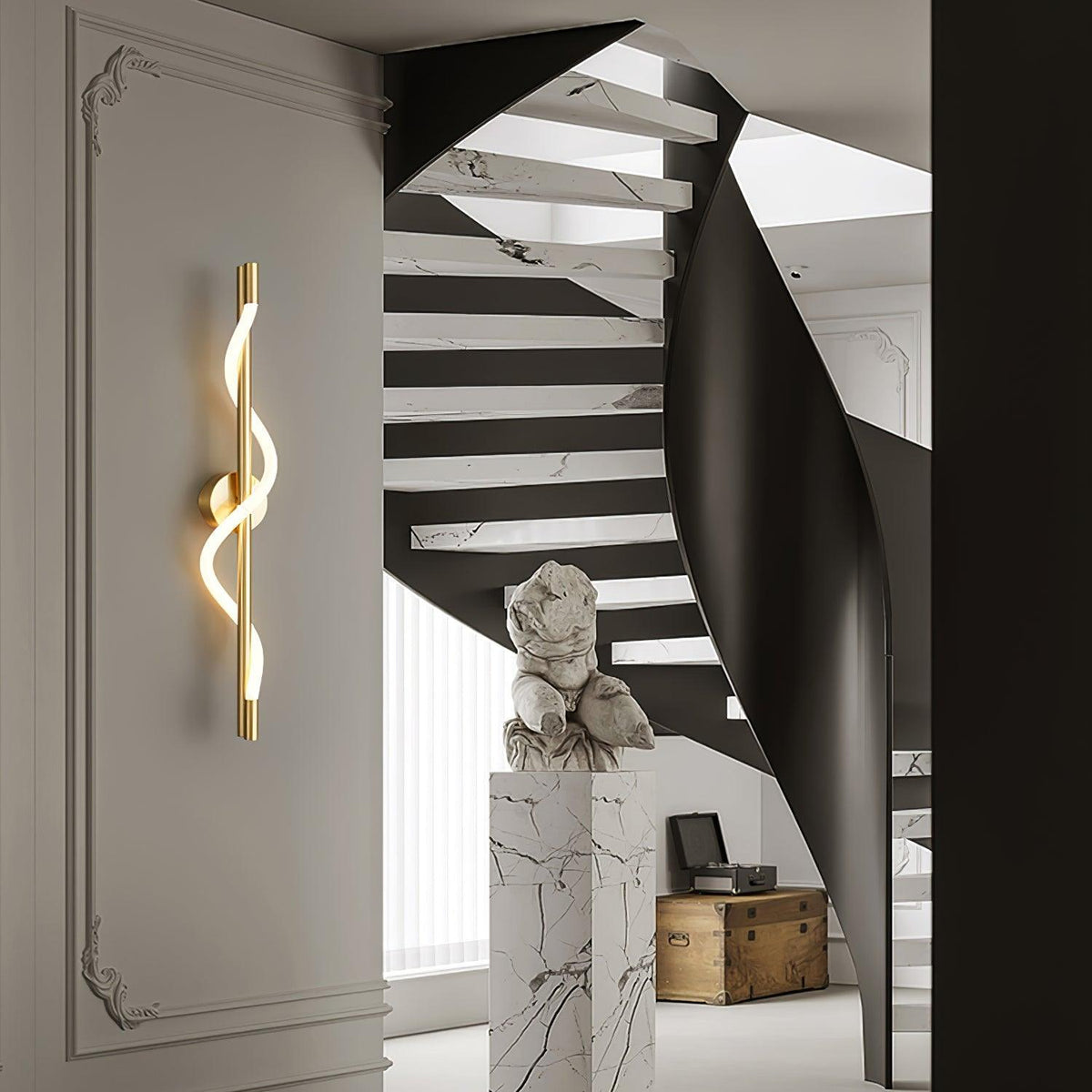 A brass Clérachic wall light with a lit spiral band hangs on a pale wall by a black curving staircase and a marble plinth.