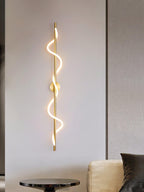 A tall brass Clérachic wall light with a lit two-turn spiral band hangs above a round side table and a cream sofa.
