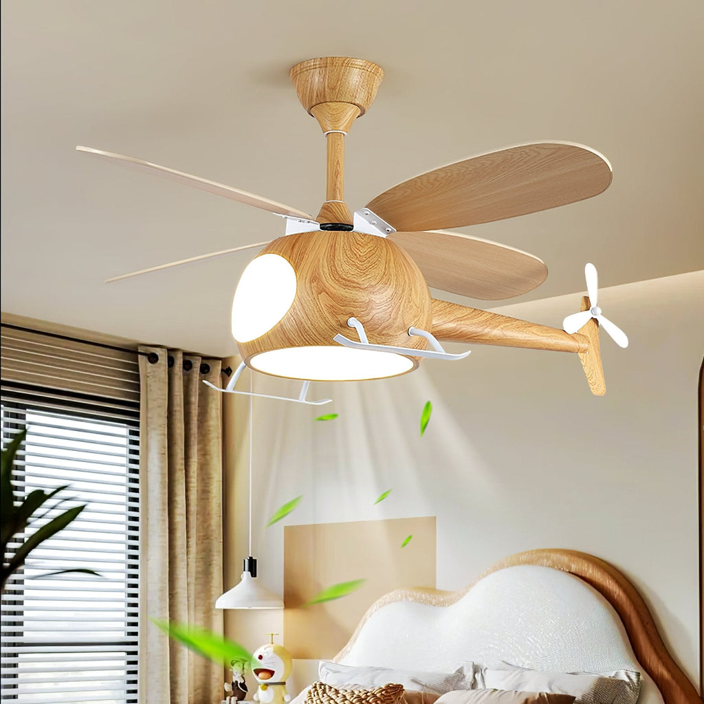 The wood-grain helicopter fan from Clérachic with its light on, blowing a stream of green leaves down towards a bed with a curved headboard.