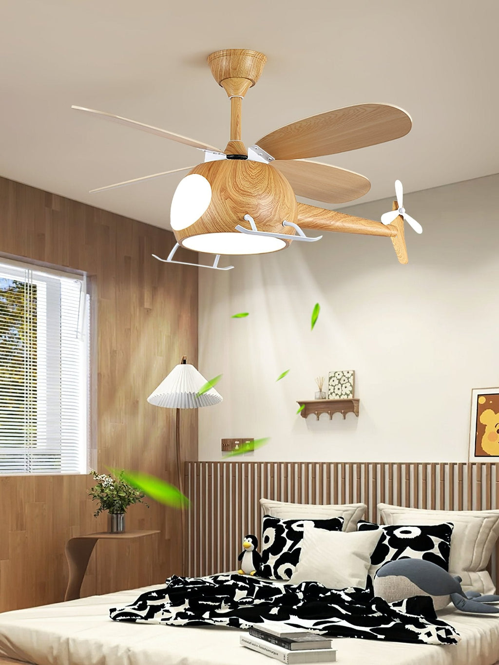 A lit wood-grain helicopter ceiling fan from Clérachic, with its blades turning above a bed, panelled wall and window, and leaves in the air.