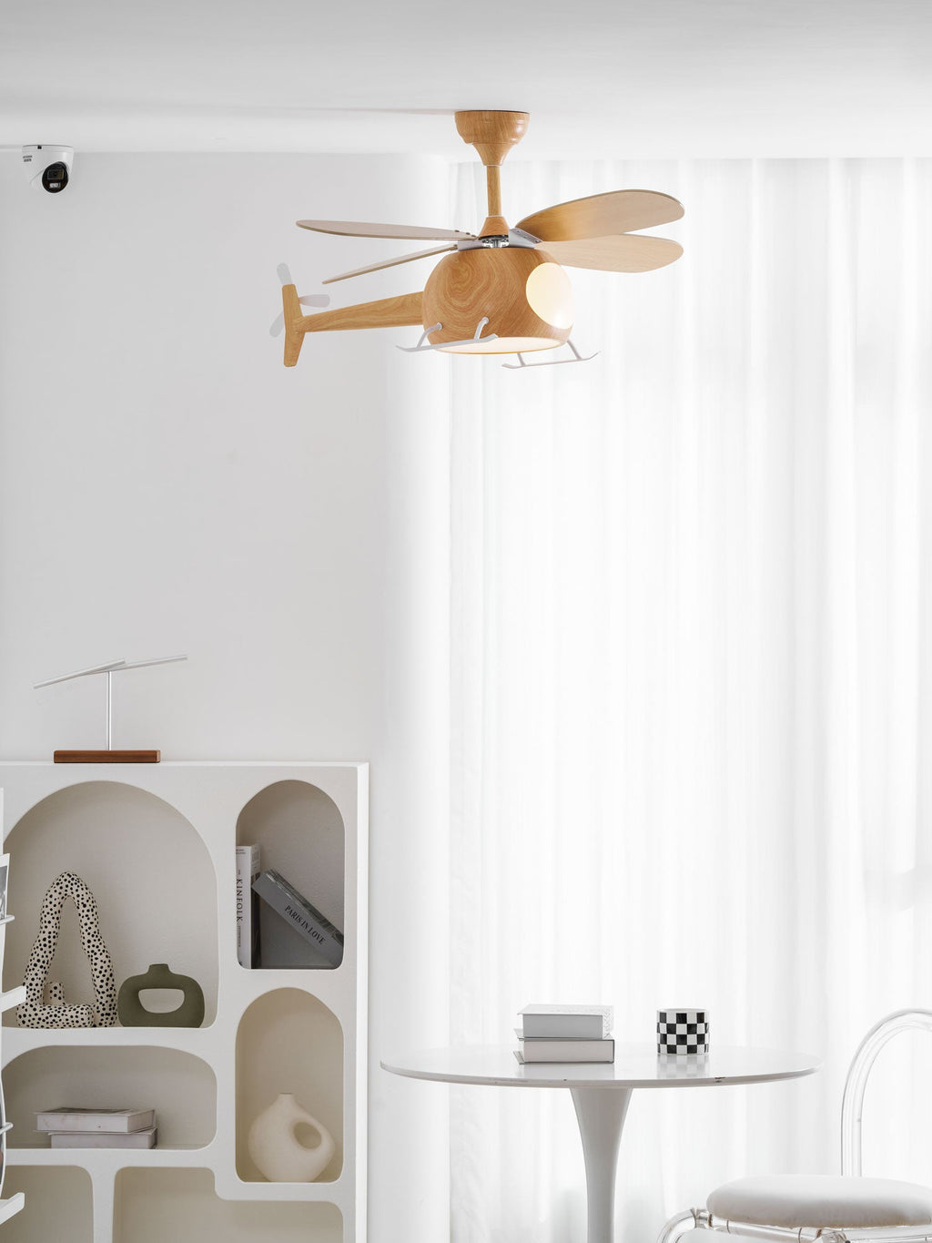 In a bright white room with sheer curtains, an arched shelf unit and a round table, a wood-grain Clérachic helicopter ceiling fan hangs overhead.
