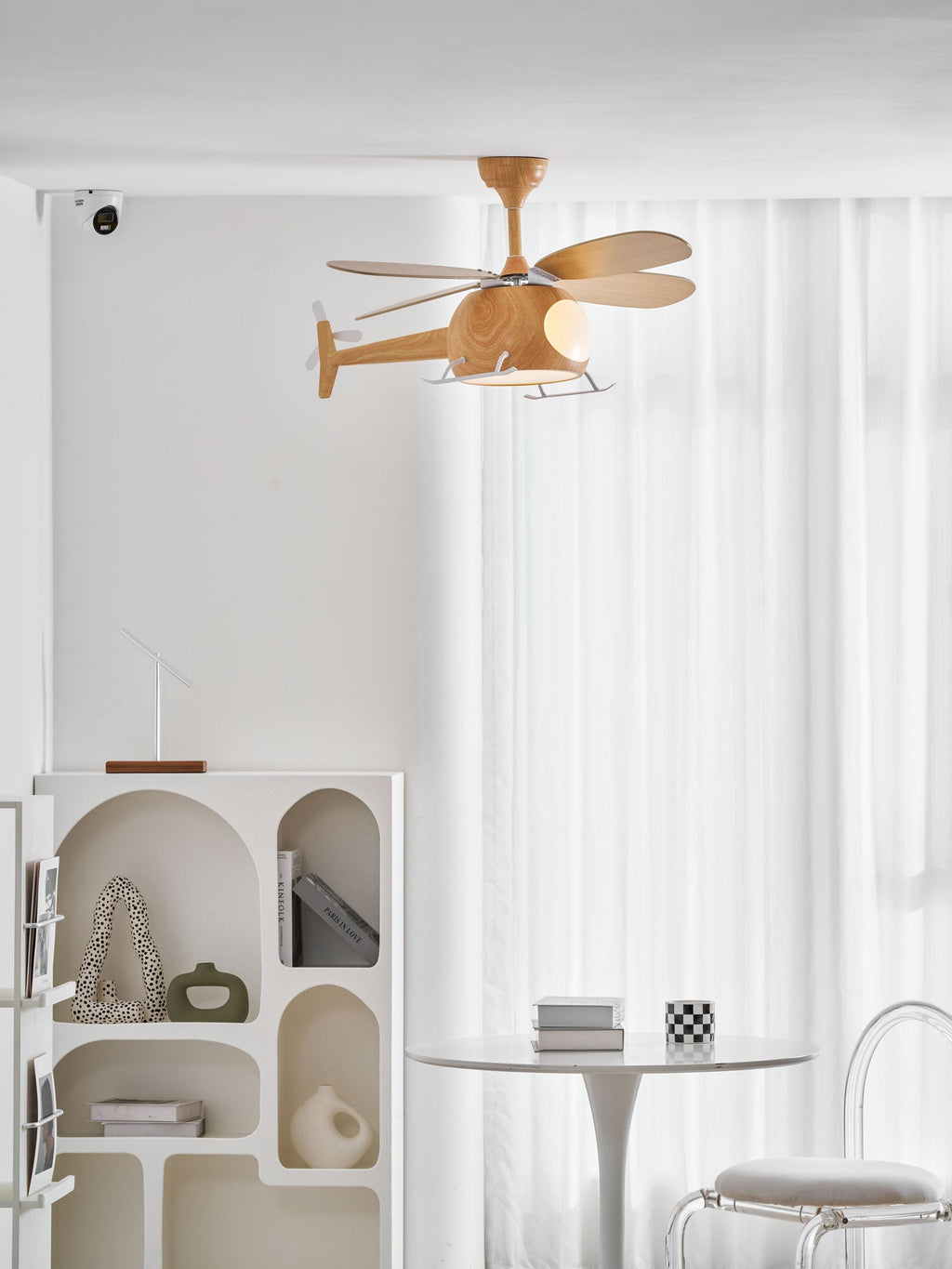 Helicopter-shaped ceiling fan from Clérachic seen from a distance in a white room, with an arched shelf unit and a white table beneath.