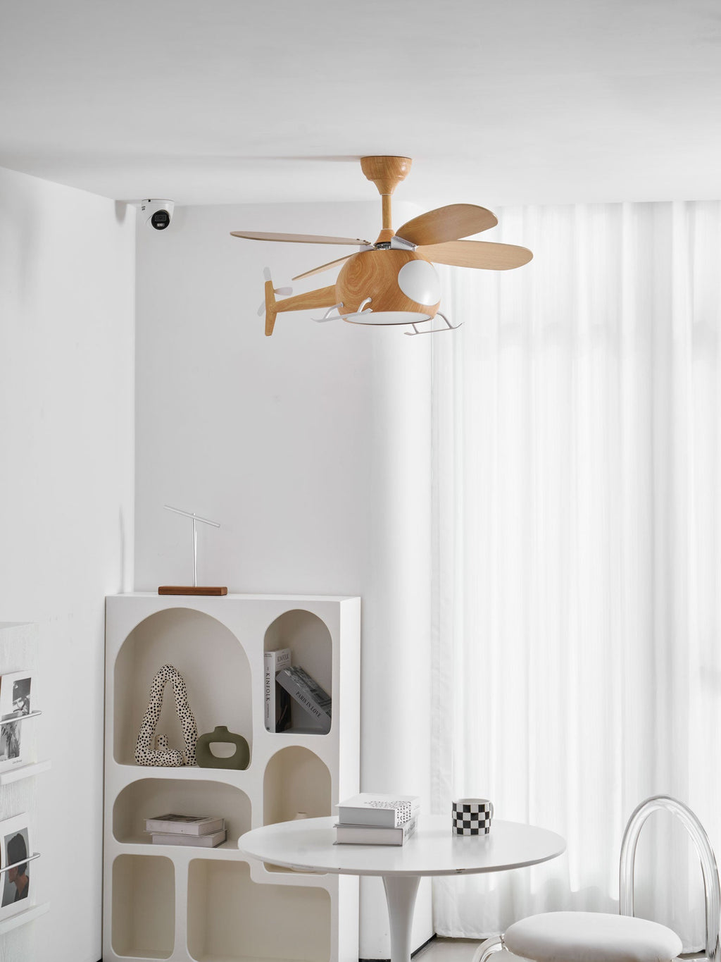 A helicopter-shaped Clérachic ceiling fan seen at a distance, with sheer curtains behind and an arched shelf and a white table below.