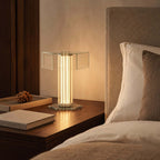 This Clérachic glass table lamp glows warm on a dark wood bedside table beside a cream headboard, a book and a small dish.