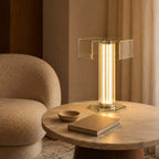 A Clérachic table lamp with a hexagonal glass top and a lit centre column stands on a round marble table beside a boucle chair.