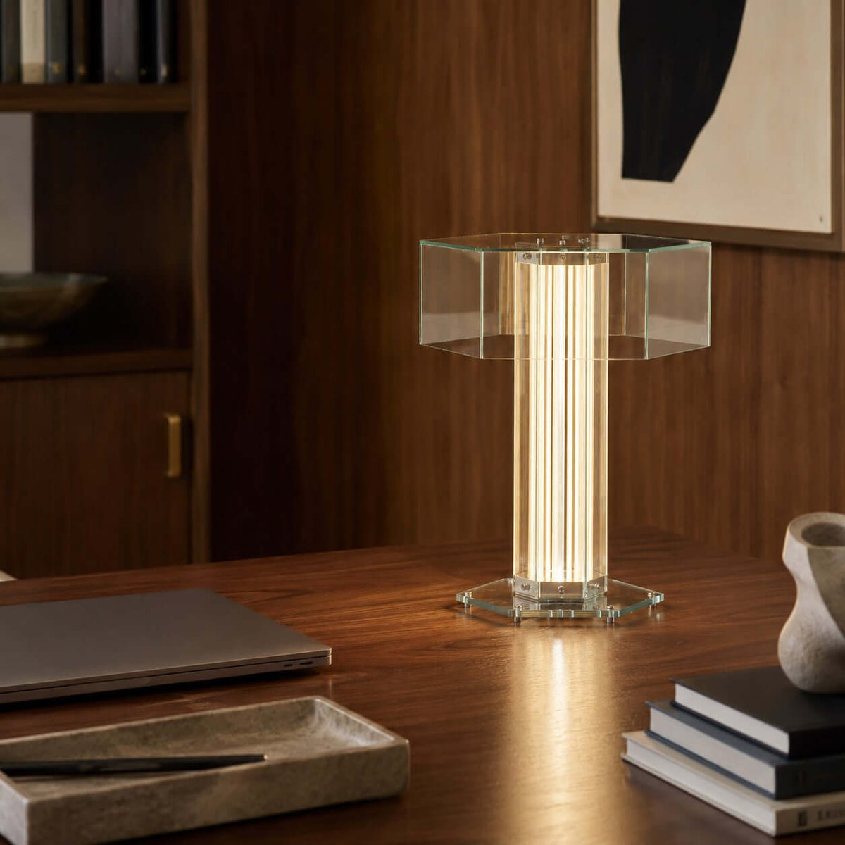 Lit Clérachic glass table lamp on a dark wooden desk, with a laptop, a stack of books and a stone tray beside it.