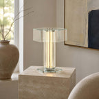 The lit column and clear glass shade of this Clérachic table lamp sit on a travertine block, beside a stone vase and a framed print.
