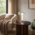 This Clérachic glass lamp sits on a dark round side table next to a cream armchair, a knitted throw and a mug.