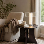 Beside a cream armchair with a brown throw, this Clérachic glass table lamp glows on a dark round table with a mug and books.