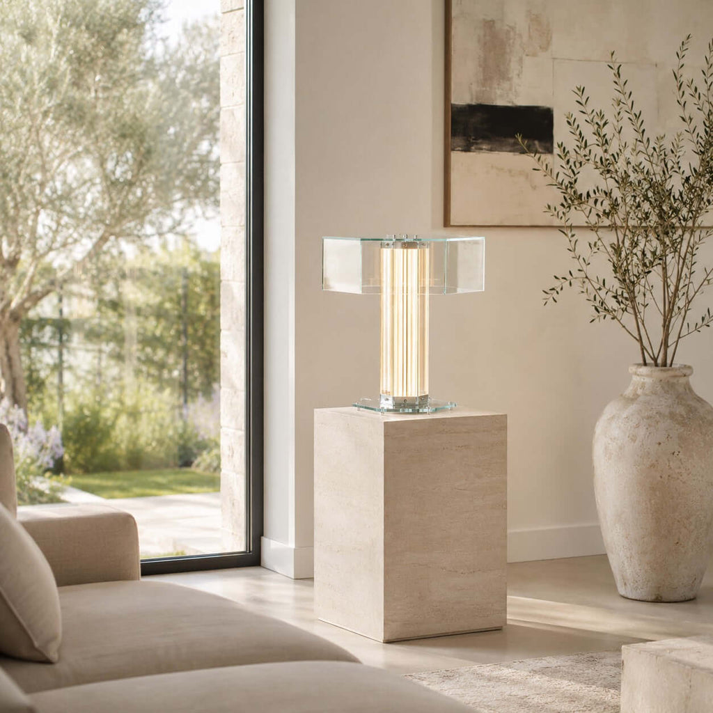 A Clérachic hexagonal glass table lamp stands lit on a travertine plinth beside a tall window and a stone vase with olive branches.