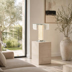 A Clérachic hexagonal glass table lamp stands lit on a travertine plinth beside a tall window and a stone vase with olive branches.