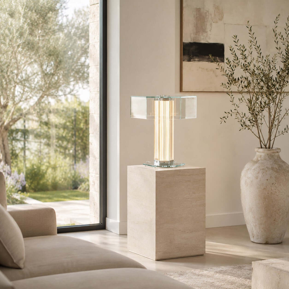 A Clérachic hexagonal glass table lamp stands lit on a travertine plinth beside a tall window and a stone vase with olive branches.