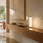 This Clérachic glass lamp glows on a wall-hung wooden shelf, next to a stone bowl and a small stack of books, in a bright hallway.