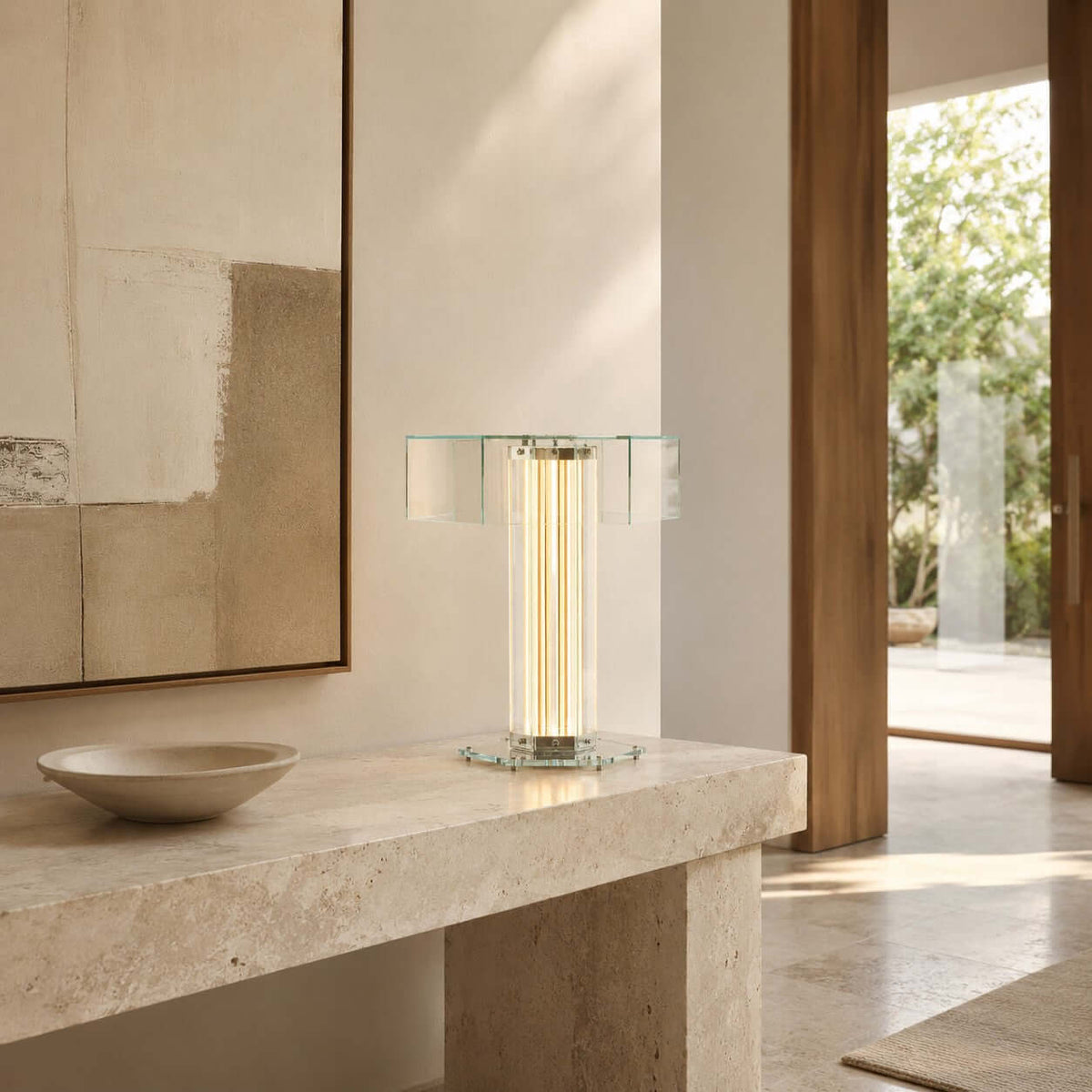 Lit Clérachic table lamp with a clear glass shade on a travertine console, beside a stone bowl, an open door and a framed print.