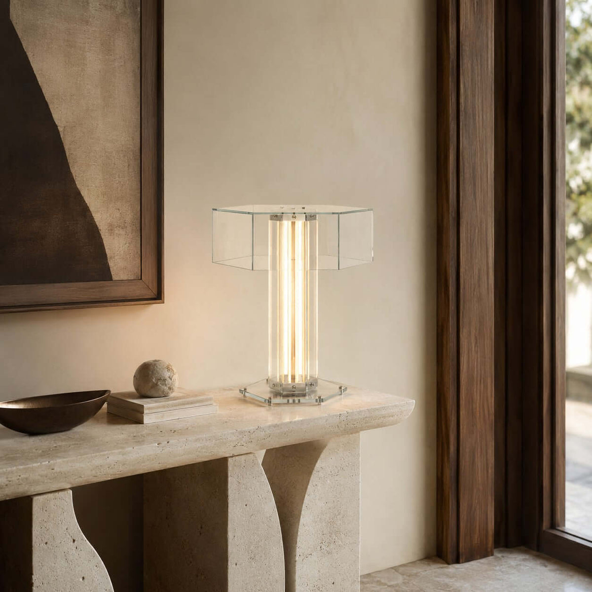 This Clérachic glass lamp stands on a curved stone console, with a dark bowl and a framed print and a wooden door frame beside it.