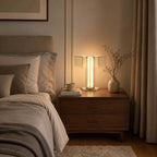 A Clérachic table lamp glows warm on a walnut bedside chest of drawers beside a bed with taupe bedding and a vase of stems.