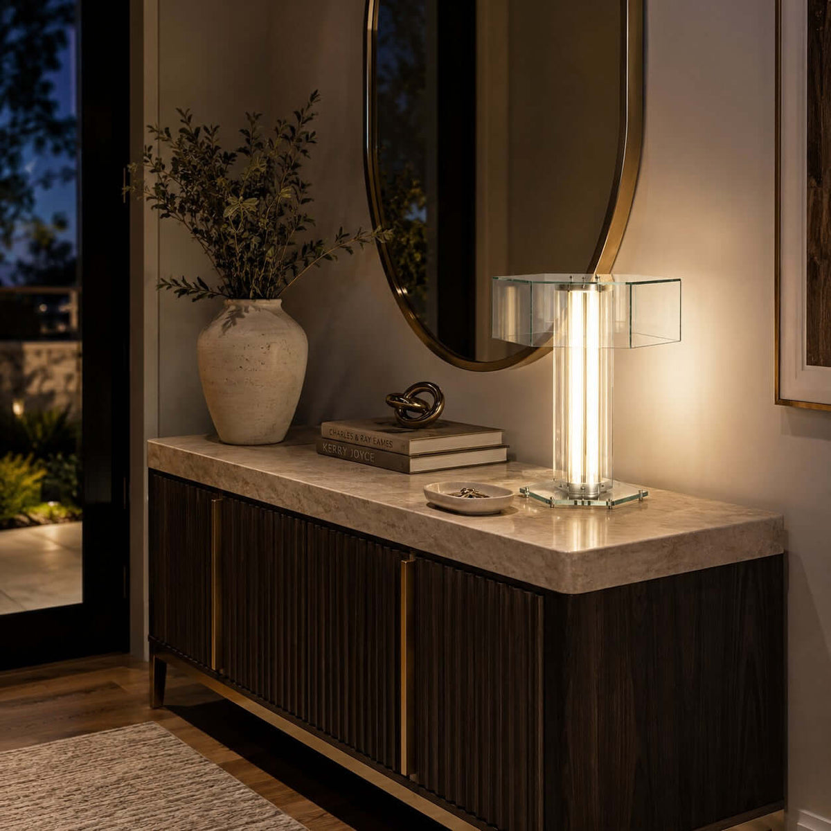 This Clérachic lamp lights up a stone-topped dark sideboard in an entrance, below a round mirror and beside a stone vase.