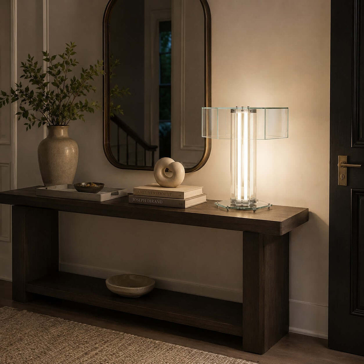 A Clérachic hexagonal glass table lamp glows on a dark wood console under an arched mirror, with a vase, books and a small sculpture.