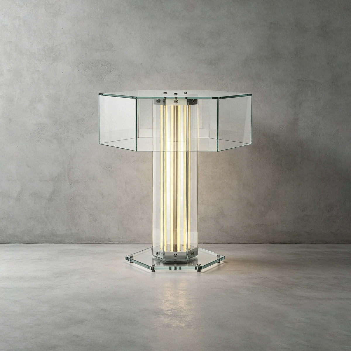The clear hexagonal glass top, glowing column and glass base of this Clérachic table lamp, against a concrete-look wall.