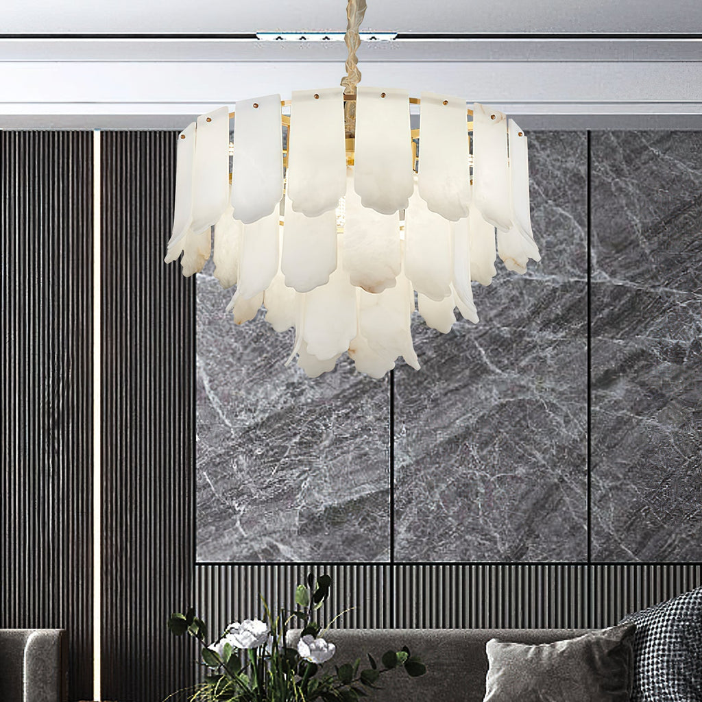 The Clérachic Hue Alabaster Chandelier lit before a grey marble feature wall, seen over a sofa and a vase of white blooms.