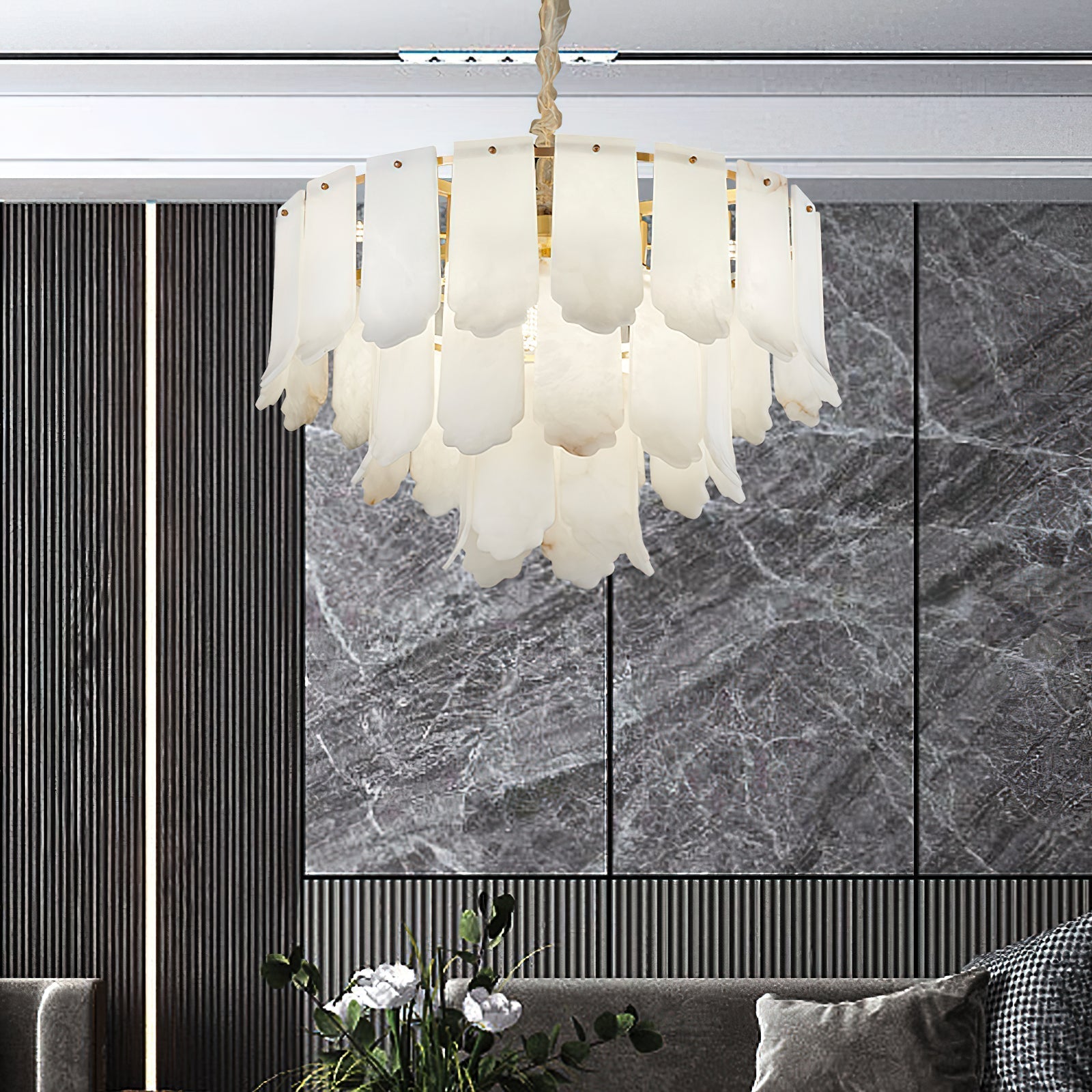 The Clérachic Hue Alabaster Chandelier lit before a grey marble feature wall, seen over a sofa and a vase of white blooms.