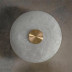 Clérachic alabaster wall sconce with a brushed brass centre cap, its veined stone disc unlit against a taupe plaster wall.