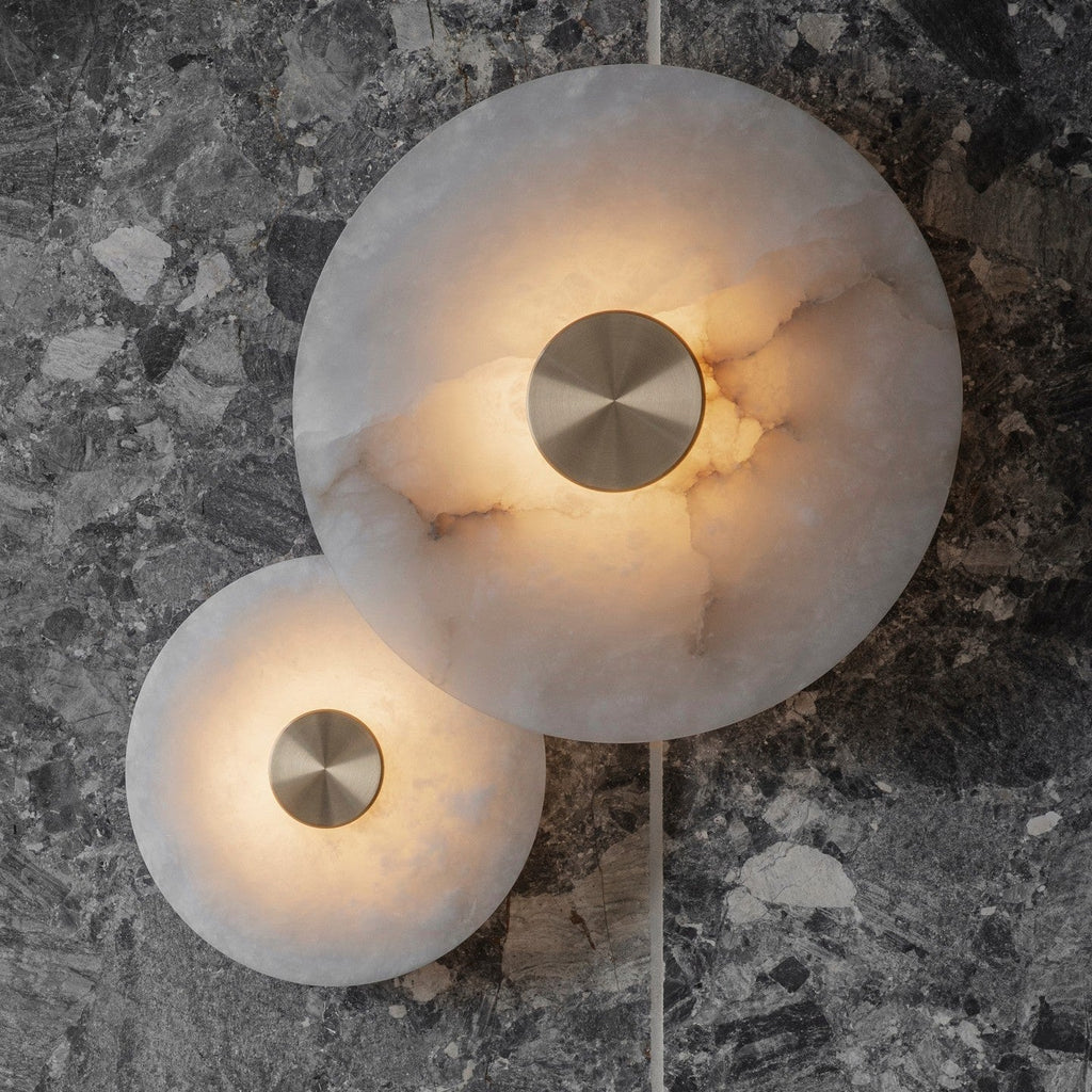 Clérachic alabaster sconces lit on a grey terrazzo wall, the stone's amber veining showing through the glow.