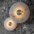 Clérachic alabaster sconces lit on a grey terrazzo wall, the stone's amber veining showing through the glow.