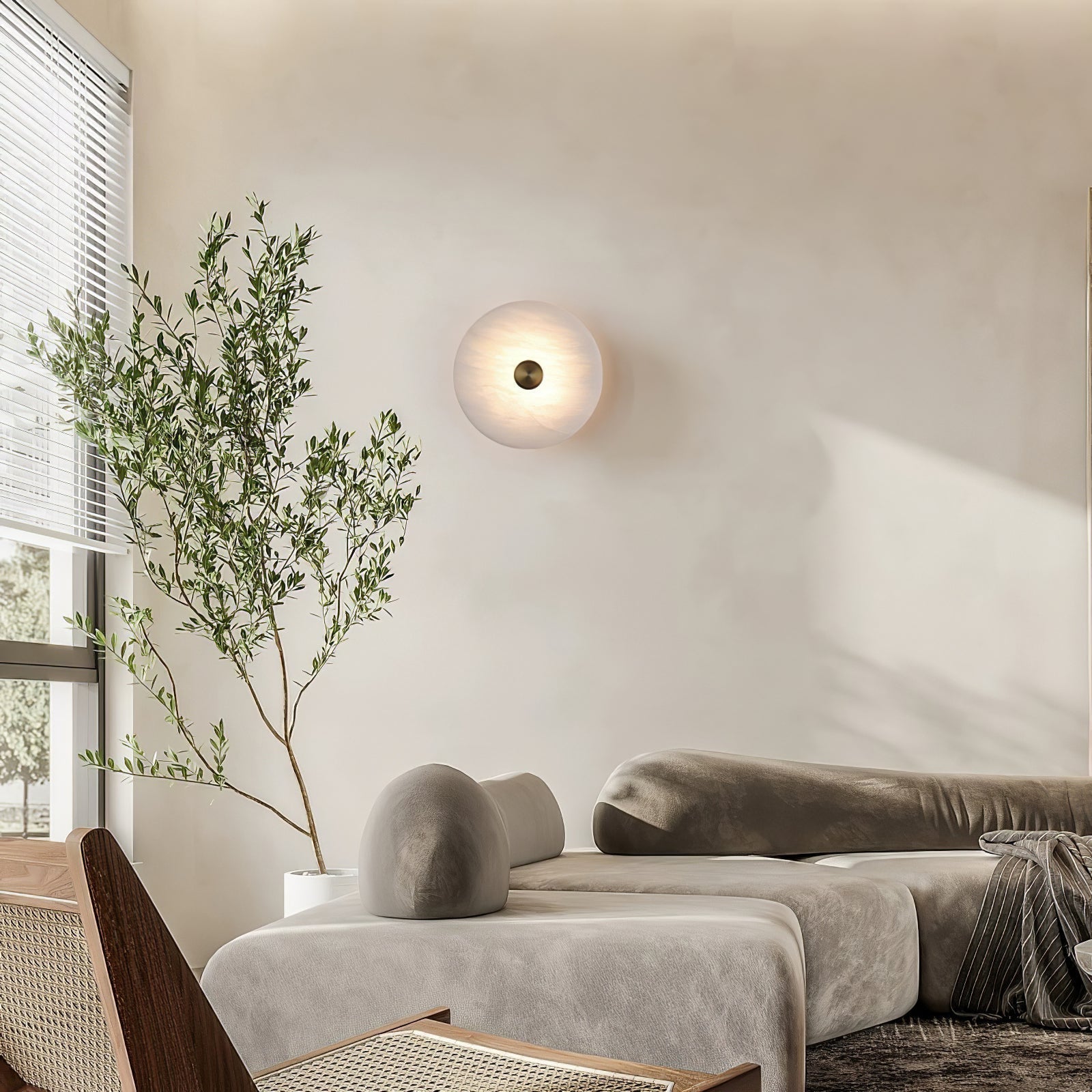 The Clérachic sconce lit above a low sofa in a bright living room, an olive tree framing the window beside it.