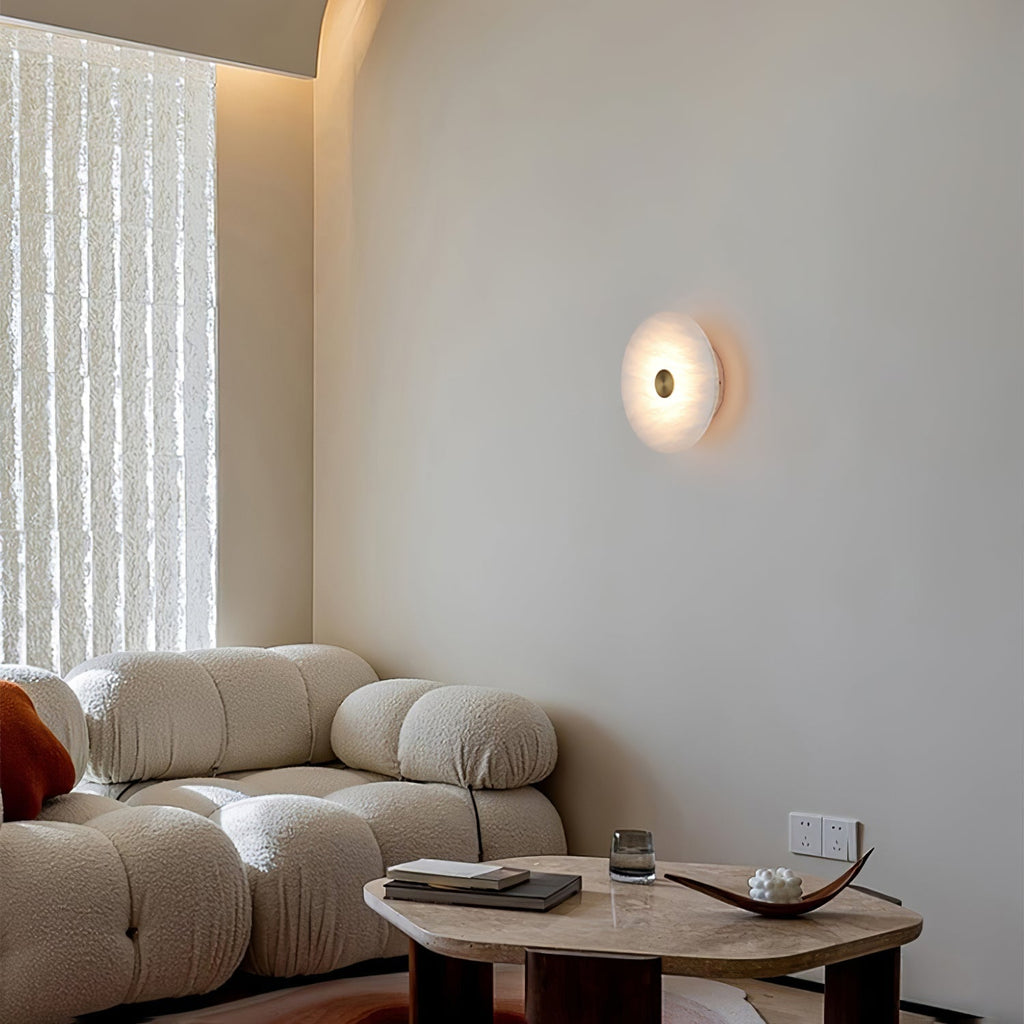 The Clérachic sconce lit on a pale wall above a bouclé sofa, beside a sheer curtain in a calm living room.