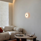 The Clérachic sconce lit on a pale wall above a bouclé sofa, beside a sheer curtain in a calm living room.