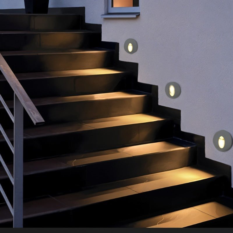 Clérachic round recessed stair lights set in a pale wall, each casting warm light across dark stone steps beside a railing.