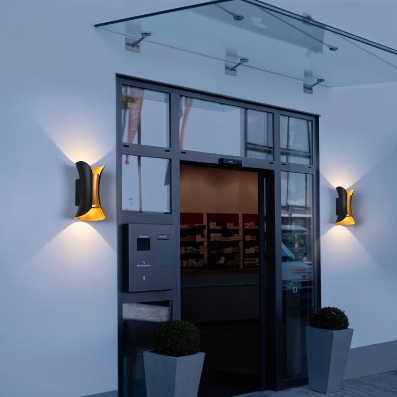 Pair of Clérachic black wall lamps with gold interiors lighting either side of a glass-canopied entrance at dusk.