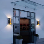 Pair of Clérachic black wall lamps with gold interiors lighting either side of a glass-canopied entrance at dusk.