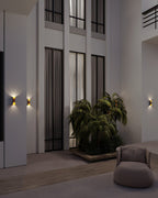 Several small Clérachic wall lamps with gold interiors glowing on pale walls in a tall hall with palms and a lounge chair.